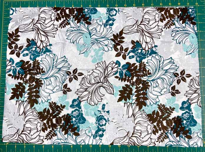 A large rectangle of grey, turquoise, silver, black and white floral fabric is shown on top of a green cutting mat. Omnigrid Triangle Ruler for Half Square Triangles, Fabric Creations Trendy Neutral Fabric collection, Fabric Creations Fabric Palette collection, Clover Hot Hemmer, UNIQUE quilting Clever Clips, UNIQUE Clear Grip, Oliso Pro TG1600 Smart Iron, Fairfield Crafter’s Choice Pillow Forms, StitchnSew EZ-Print Quilt Block Sheets