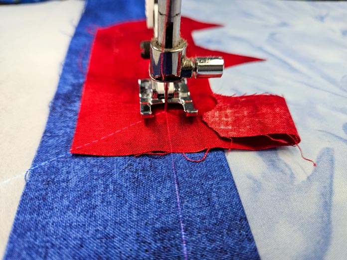 An open-toe sewing machine foot with the needle in the down position is threaded through a red piece of fabric placed on top of a blue fabric strip. The machine's stitch projection is turned on. Husqvarna VIKING DESIGNER EPIC 3 sewing and embroidery machine, Husqvarna VIKING 6in/15.2cm Applique Scissor, Husqvarna VIKING Open Toe Presser Foot for IDF System, SULKY Slimline Metallic Dream Assortment