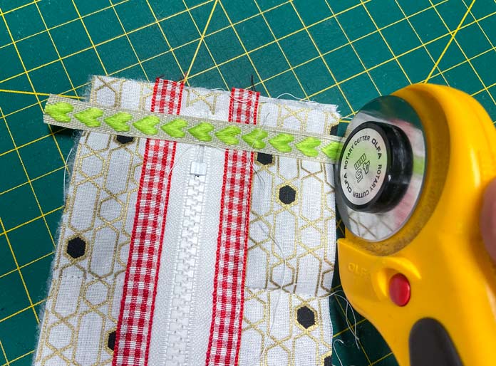 A yellow rotary cutter trims off the ends of a ribbon with green hearts sewn across the end of a zipper on a small quilt block. A green cutting mat is in the background. UNIQUE SEWING Self-Gripping Fastener Strip - 25cm (10″) - White, UNIQUE Webbing 25mm x 10m (1″ x 11yd) - White, ELAN Trench Buckle - 45mm (13⁄4″) - Brown, UNIQUE CREATIV Trim Pack - Black & White, UNIQUE SEWING Invisible Magnetic Snaps - Sew-On - 12mm (1⁄2″) - 2 sets,  SOFTKUT Pinking Shears (Scissors) - 81⁄2″ (21.6cm), COSTUMAKERS General Purpose Closed End Zipper 20cm (8″) - White - 1700, UNIQUE CREATIV Trim Pack - Red & Green, OLFA RTY-2/DX - Deluxe Ergonomic Handle Rotary Cutter 45mm, UNIQUE QUILTING Clever Clips Small - 12 pcs, UNIQUE SEWING Double Adjust Parachute Buckle - Plastic - 25mm (1″) - White, CRAFTING ESSENTIALS Bottle of Buttons - Pink Tones - 75g (2.6oz), UNIQUE Braided Elastic 3mm x 3.7m - White, free tutorial