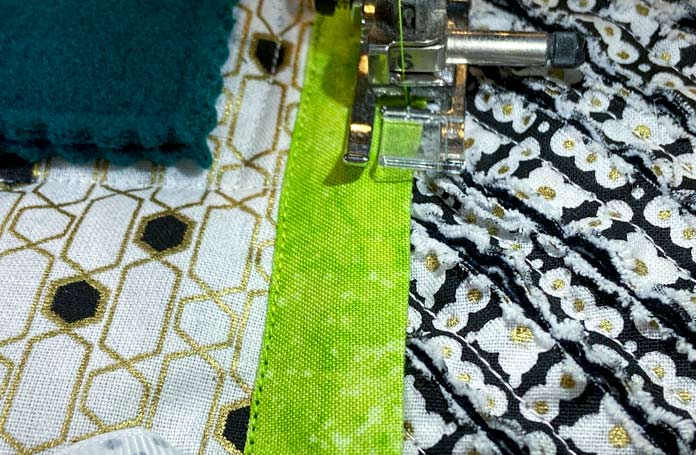 A sewing machine stitches along the edge of a lime green strip of fabric. Black, white, and gold fabrics are visible on either side of the green strip while a dark green square of wool felt can also be seen in the top corner of the photo. UNIQUE SEWING Invisible Magnetic Snaps - Sew-On - 12mm (1⁄2″) - 2 sets, COSTUMAKERS General Purpose Closed End Zipper 20cm (8″) - White - 1700, UNIQUE SEWING Double Adjust Parachute Buckle - Plastic - 25mm (1″) – White, UNIQUE QUILTING Clever Clips Small - 12 pcs, CLOVER 7812 Roll & Press, UNIQUE QUILTING Wool Pressing Mat - 18″ x 24″ - Grey, OLISO M2Pro Mini Project Iron with Solemate, CLOVER 240 - Fork Pins - .56mm, CLOVER 464/18 - Bias Tape Maker - 18mm (3⁄4″), Odif 505 Temporary Quilt Basting Adhesive Fabric Spray - 156g, FAIRFIELD Low-Loft® Quilt Batting - Crib - 114 x 152cm (45″ x 60″), free tutorial