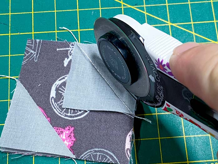 A hand uses a white rotary cutter to trim away the excess fabric on a brown square of fabric that has two gray squares of fabric sewn to it. A green cutting mat is in the background.