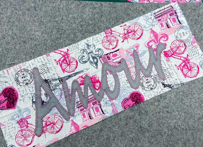 A rectangular piece of pink, gray and white fabric with the word amour appliqued on top sits on top of a gray wool pressing mat.