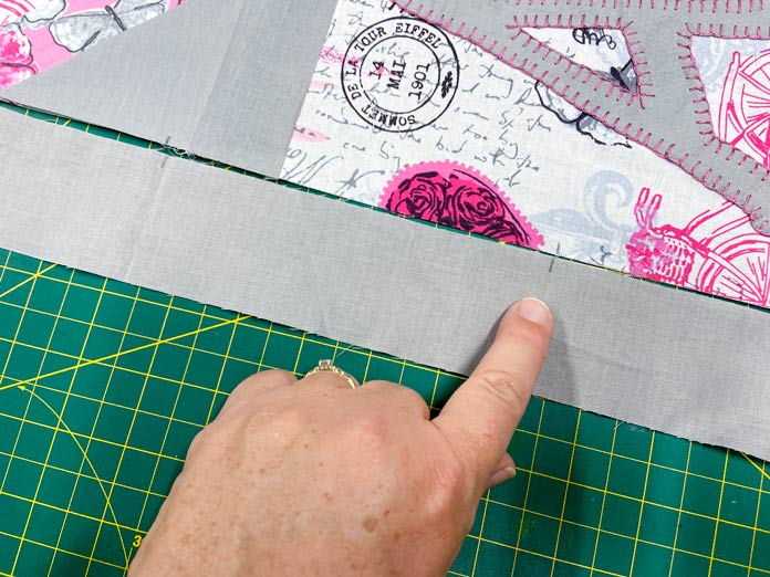 A hand points to small pencil marks along the edge of a pink, white and gray quilt top and a gray border. A green cutting mat is in the background. 