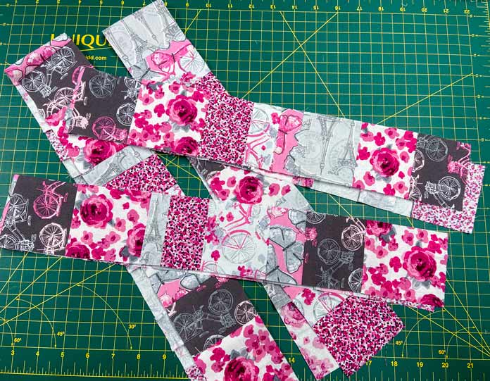 Four long strips of pieced fabrics lie on top of a cutting board. The strips are made up of fabrics that are prints of pink, white, gray and maroon.