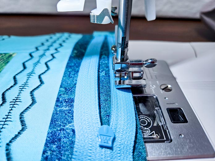 A blue zipper on blue fabric under a metal presser foot on a sewing machine