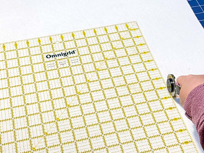 A hand is shown using a rotary cutter and a large square rotary cutting ruler to cut a square of white interfacing.