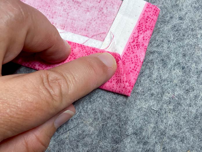 A hand is shown holding down the folded edge of the end of a strip of pink fabric, so that it encases the white interfacing neatly inside. Everything sits on a grey pressing mat.