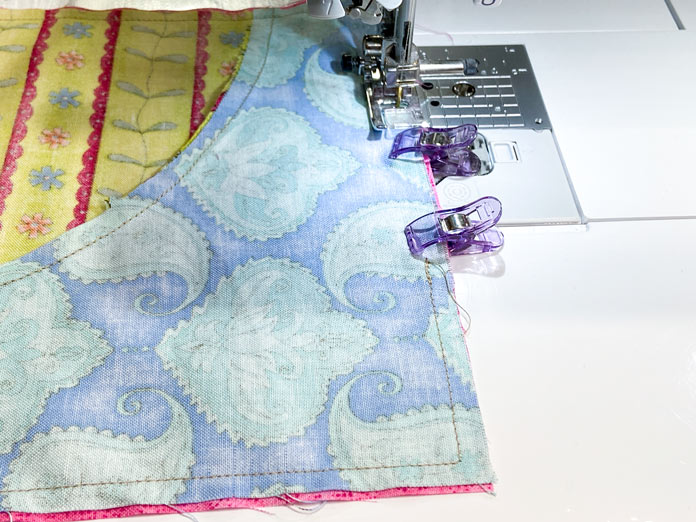 A sewing machine sews along all four sides of a quilt block that has been clipped to the same-sized square of fabric.