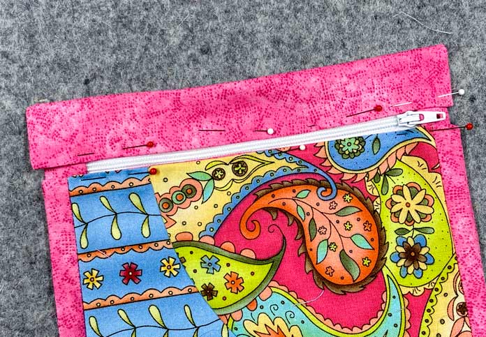A multicolored pocket and a pink rectangle of fabric are pinned to a white zipper. Everything sits on a grey pressing mat.