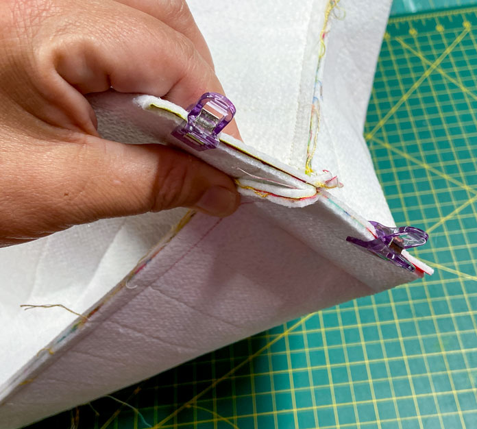 A hand is shown holding the corner of a tote bag. Purple clips hold the seam together. A green cutting mat is visible in the background. 