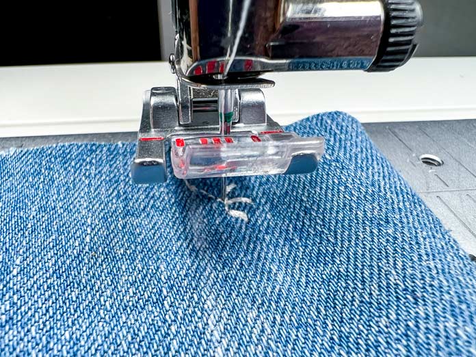 Close-up of the 2A Fancy Stitch Foot on the PFAFF Creative Icon 2 sewing machine during the ribbon stitch process. The machine has completed the initial short section of stitching and paused automatically, as part of the programmed sequence. The white decorative thread is visible on the denim fabric, aligned with the center guide of the presser foot, ready for the next step.