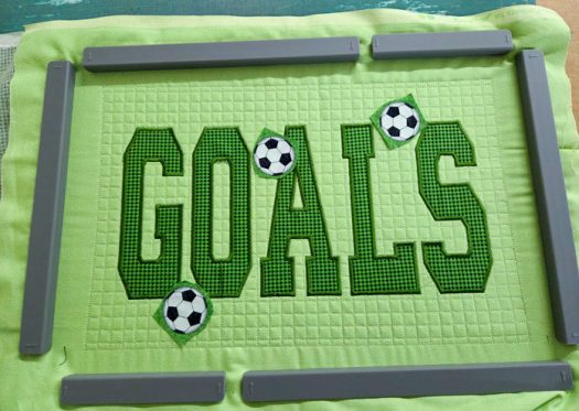 The word GOALS on green fabric with three fabric soccer balls