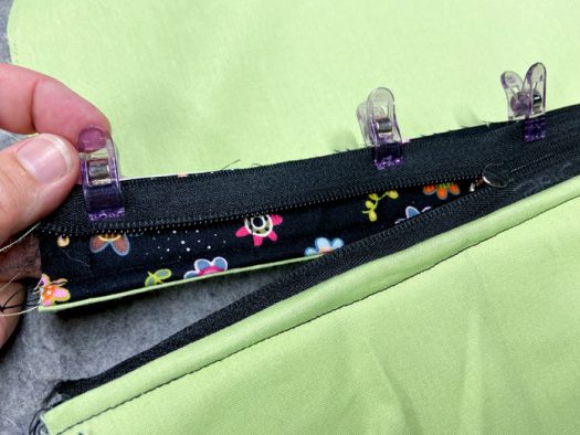 A hand holds a black zipper that is clipped to a piece of black floral fabric. Green lining fabric is visible on both sides of the zipper. This sits on top of a grey pressing mat.