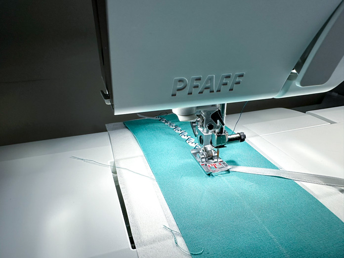 A light green 4” x 12” piece of fabric with a partially stitched, folded ¼” ribbon, in the process of being stitched down using the single ribbon stitch, stopped to show ribbon under the foot.