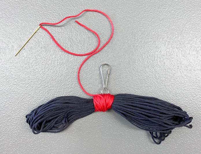 Red floss wrapped around a black skein with a clip attached in the middle, and a length of loose floss threaded in an embroidery needle; DMC Matte Cotton Yarn and Elan Snap-Clip
