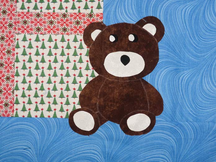 A brown and white fabric teddy bear fused to the quilt top of a Christmas-themed fabric wall hanging; Odif 808 Spray and Fix Temporary Adhesive for Paper Patterns, Odif 606 Spray and Fix No-sew Fusible Adhesive Web, Odif 404 Spray and Fix Permanent Repositionable Adhesive for Craft Material, Odif 505 Temporary Quilt Basting Adhesive Fabric Spray