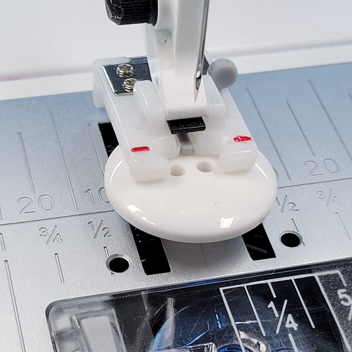 A button sitting in the slot of a presser foot used to sew a button with the sewing machine; Husqvarna Viking Designer Ruby 90, Button Foot with Placement Tool