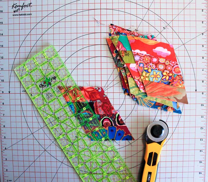 Aligning your ruler to cut two 1½” strips from the step 4 units shows a quilting ruler, a rotary cutter, and a stack of pieced units on a cutting mat; Komfort Kut 360° Rotating Cutting Mat - 18" x 18", Sew Easy Triangle Ruler 60°, OLFA Quick Change Rotary Cutter