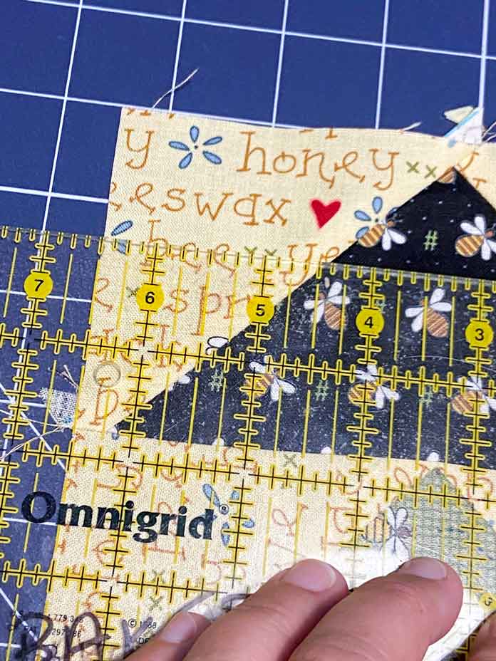 A hand is shown holding a quilting ruler on top of a black and yellow flying geese block. The ruler shows that the block is 6½" wide; Omnigrid ruler - 6" x 24", OLFA Double Sided Cutting Mat (navy blue)