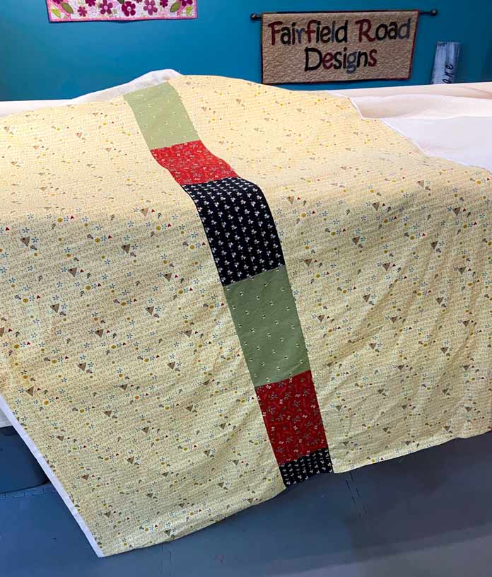 A backing made of yellow fabric with a band of black, red and green fabrics down the center lays on the bed of a long-arm quilting machine.
