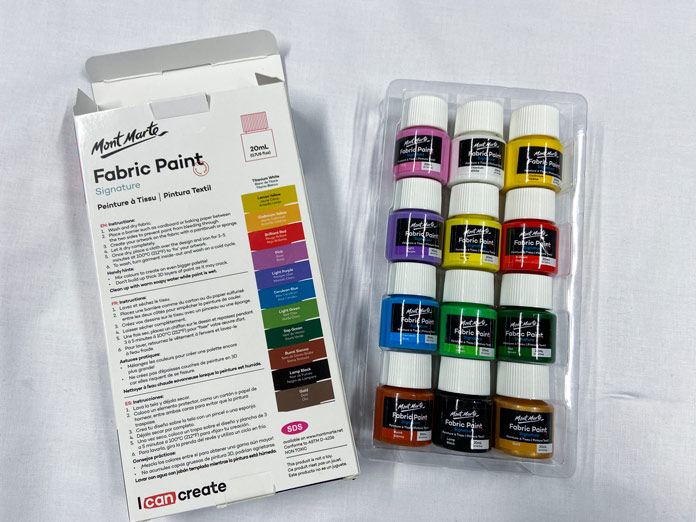A white box and a clear tray holding 12 small tubs of multicolored paints sit on a white background; Mont Marte Signature Fabric Paints