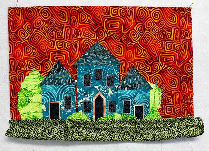 3 blue houses, green bushes and trees are placed on an orange print background with green fabric at the bottom for grass