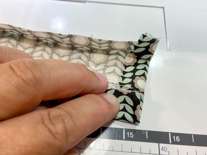 A hand is shown using a sewing machine to topstitch along the folded edge of the fabric on a purse strap. A purple clip holds the folded fabric in place. 