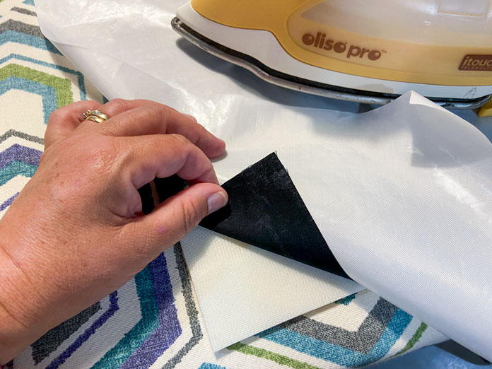 A hand is shown folding back a corner of a piece of black fabric to expose the fusible foam underneath. A pressing sheet sits on top of the black fabric and a yellow iron sits on top of the pressing sheet.