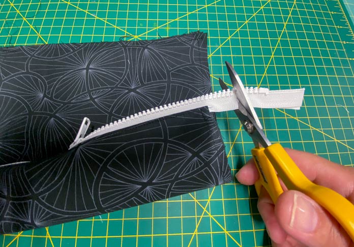 A hand is shown using yellow scissors to cut off the end of a white zipper. A green cutting board is in the background. 