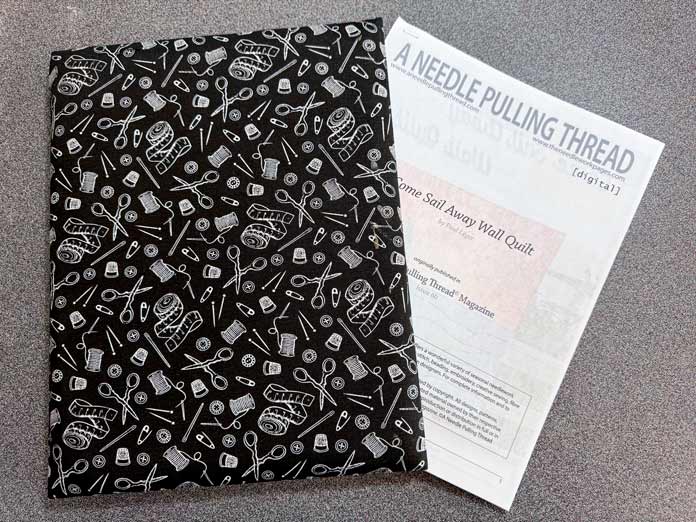 A Hemline Gold Quilters 4-in-1 Multi-Mat of a black and white sewing print with a printed digital pattern from A NEEDLE PULLING THREAD partially inserted under the front cover. All sit on a gray table. Hemline Gold Multi-Use Craft Bag (28.5cm x 36.5cm x 17.8cm), Hemline Gold Sewing Kit, Hemline Gold Quilters 4-in-1 Multi-Mat (30cm x 24cm), Hemline Gold Quilters Clips (Pack of 30), Hemline Gold Retractable Tape Measure - 150cm/60, oliso M3Pro Project Iron – Coral, OLFA 45mm RTY-2/DX/MAG Ergonomic Rotary Cutter – Magenta, OLFA RM-MG - 24″ x 36″ Double Sided Rotary Mat, UNIQUE Sewing Tools Holder, OLFA QR-6x12 - 6″ x 12″ Frosted Acrylic Ruler, OLFA QR-6x24 - 6″ x 24″ Frosted Acrylic Ruler, Fairfield Quilter's 80/20 Quilt Batting - 229 x 274cm (90″ x 108″) 