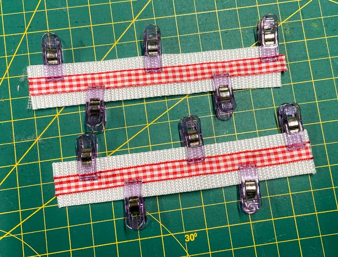 Small purple clips hold pieces of white webbing and red gingham ribbon. A green cutting mat can be seen in the background. UNIQUE SEWING Self-Gripping Fastener Strip - 25cm (10″) - White, UNIQUE Webbing 25mm x 10m (1″ x 11yd) - White, ELAN Trench Buckle - 45mm (13⁄4″) - Brown, UNIQUE CREATIV Trim Pack - Black & White, UNIQUE SEWING Invisible Magnetic Snaps - Sew-On - 12mm (1⁄2″) - 2 sets,  SOFTKUT Pinking Shears (Scissors) - 81⁄2″ (21.6cm), COSTUMAKERS General Purpose Closed End Zipper 20cm (8″) - White - 1700, UNIQUE CREATIV Trim Pack - Red & Green, OLFA RTY-2/DX - Deluxe Ergonomic Handle Rotary Cutter 45mm, UNIQUE QUILTING Clever Clips Small - 12 pcs, UNIQUE SEWING Double Adjust Parachute Buckle - Plastic - 25mm (1″) - White, CRAFTING ESSENTIALS Bottle of Buttons - Pink Tones - 75g (2.6oz), UNIQUE Braided Elastic 3mm x 3.7m - White, free tutorial