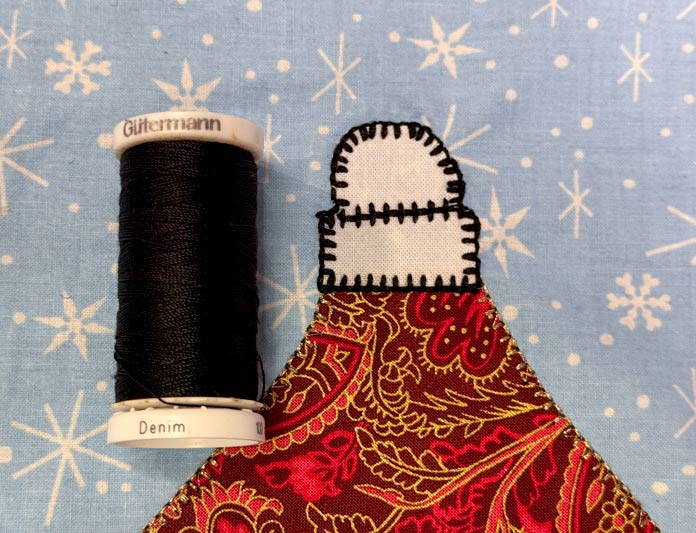 A spool of black Gütermann denim thread sits beside the top of a completed applique Christmas ornament of red fabric with a white top on light blue snowflake fabric; Gütermann Dekor Metallic Thread 200m – Gold, Gütermann Dekor Metallic Thread 200m - Col. 41, Gütermann Dekor Metallic Thread 200m – Red, Gütermann Dekor Metallic Thread 200m - Christmas Green, Gütermann Dekor Metallic Thread 200m - Col. 1005, Gütermann Denim Thread 100m – Black, UNIQUE Baby Rick Rack 3mm x 3.7m – Scarlet, UNIQUE Medium Rick Rack 6mm x 2.3m - Emerald Green, UNIQUE Jumbo Rick Rack 10mm x 2.3m - Metallic Gold, SCHMETZ #1743 Metallic Needles Carded - 80/12 - 5 count, SCHMETZ #1752 Metallic Needles Carded - 90/14 - 5 count, LDH 8½“ Craft Scissors – Purple, HeatnBond Lite Iron-On Adhesive - 43cm x 30m (17″ x 32 ¾yds) bolt, HeatnBond Stitch n Sew Tear-Away Stabilizer - 50.8cm x 22.8m (20″ x 25yds), oliso M3Pro Project Iron – Coral, OLFA 45mm RTY-2/DX/MAG Ergonomic Rotary Cutter – Magenta, UNIQUE Quilting Wool Pressing Mat - 18″ x 24″ - Grey