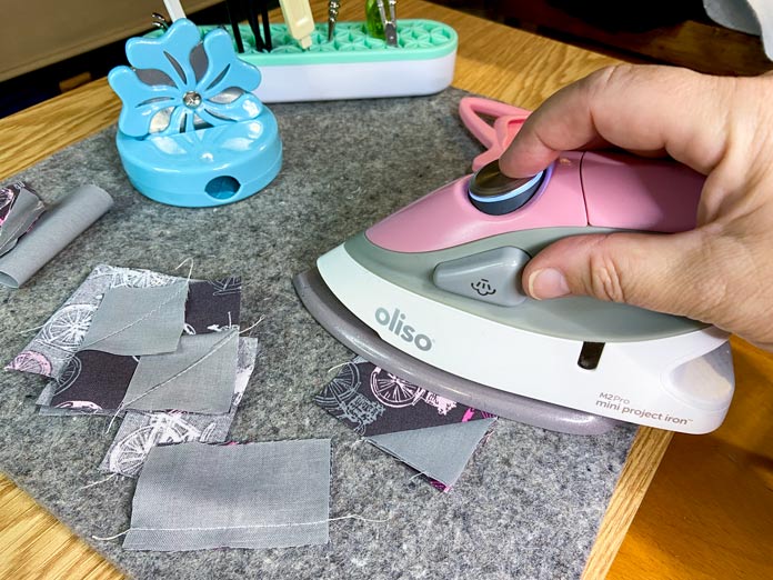 A hand is shown pressing a pieced square with a small pink iron on a wool pressing mat. 