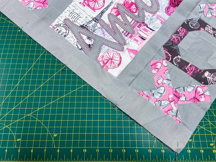 A pink, white and gray quilt top with a gray border pinned to its edge sits on top of a green cutting mat.