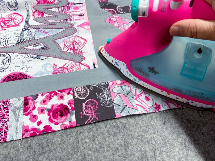 A pink iron presses the seam of a scrappy border made with pink, white, gray and maroon printed fabrics. A mottled gray wool pressing mat in is the background.