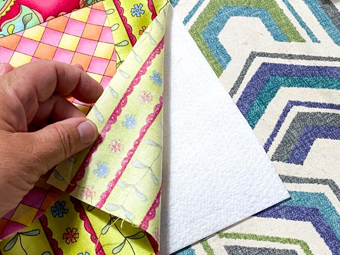 A hand is shown folding back the fabric of the block to show the square of fusible interfacing underneath. A multicolored ironing board cover is in the background.