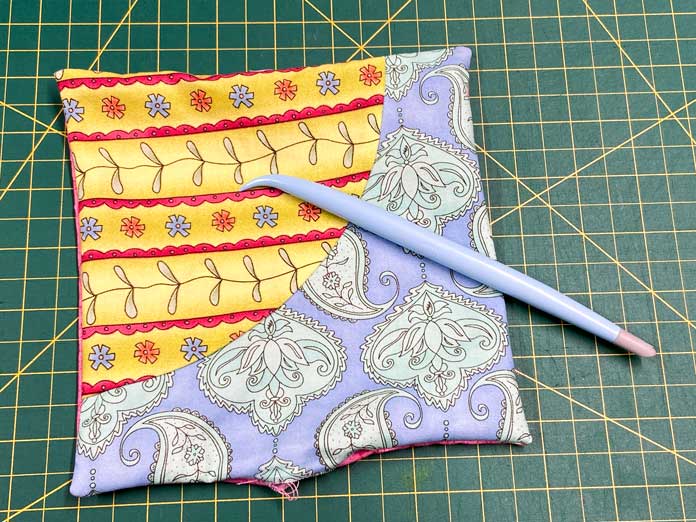 A blue-handled stiletto with a curved point sits atop a yellow and blue quilt block. A green cutting mat is in the background.