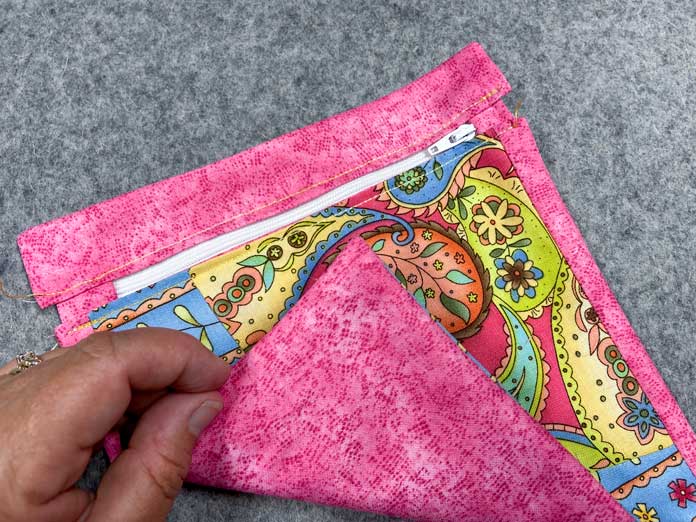 A hand folds over one corner of a multicolored zippered pocket to show that there are no raw edges on the backside. The pocket sits on a grey pressing mat.