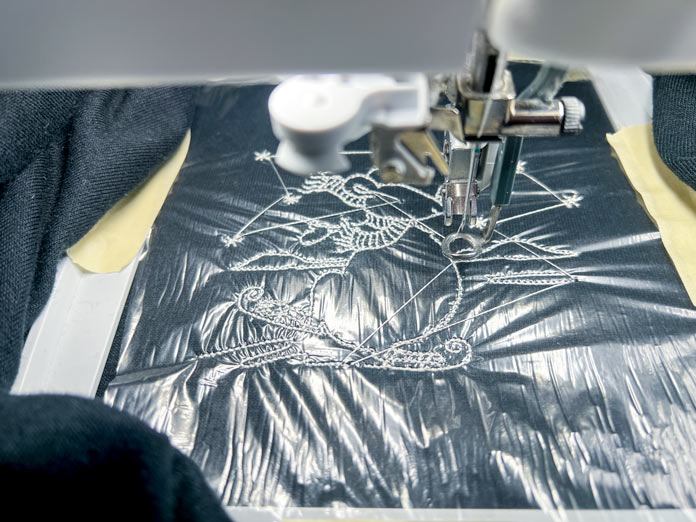 Design is being stitched in white 40wt polyester thread using the PFAFF 6D Dynamic Spring Foot. PFAFF Aqua Magic Stabilizer or pellon Sol-U-Film is applied over the fabric and secured with masking tape. The hoop is attached to the embroidery unit of the PFAFF creative icon 2.