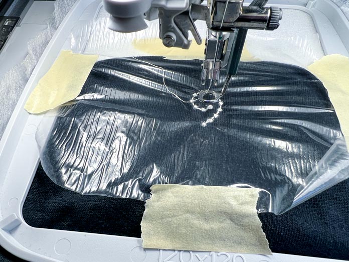 The sleeve is hooped in the PFAFF creative 120 Square Hoop (120 x 120 mm) with PFAFF Cut-a-Way Light Stabilizer and topped with PFAFF Aqua Magic Stabilizer (or Pellon Sol-U-Film), secured with masking tape. A 40wt polyester thread is being stitched using the PFAFF 6D Dynamic Spring Foot, with the machine needle working precisely through the stabilizer layers.