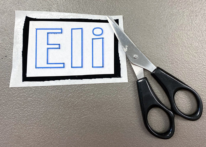 The name Eli printed on white paper placed on a piece of black fabric fused to a piece of HeatnBond Lite Iron-On Adhesive, and a pair of scissors.