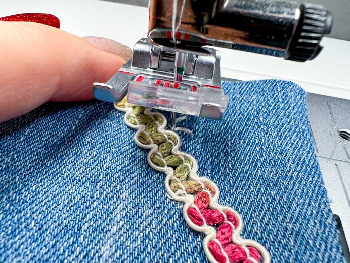 A hand carefully places variegated ribbon in front of the needle, guiding it at an angle to match the direction of the previous stitches. The 2A Fancy Stitch Foot is attached and the machine is paused, ready to resume the next section of a single ribbon stitch on blue denim fabric. 