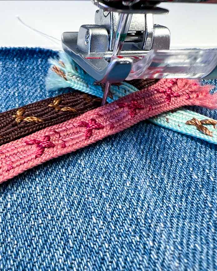 Close-up of the 2A Fancy Stitch Foot on the PFAFF creative icon 2, with brown, teal and pink ribbons positioned on a denim base. The machine is paused mid-stitch, and the third (pink) ribbon is placed in front of the needle, crossing over the second (teal) ribbon and parallel the first ribbon (brown), ready for the next step in the Triple Ribbon Stitch.