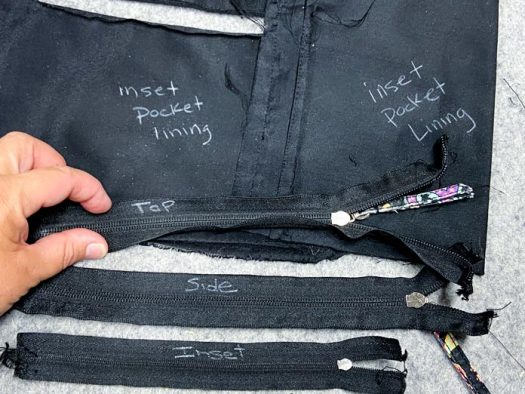 A hand holds one black zipper that has been marked “top: beside two other shorter zippers. Two pieces of black lining fabric also lie on top of a grey pressing mat.