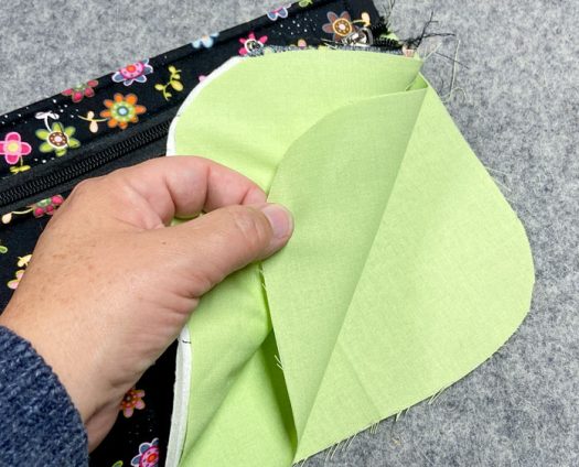 A hand holds three pieces of lining sewn to a zipper, forming a pocket. Everything sits on top of a grey pressing mat.