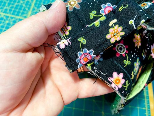 A hand is shown holding the corner of a tote bag. Purple clips hold the seam together. A green cutting mat is visible in the background. 