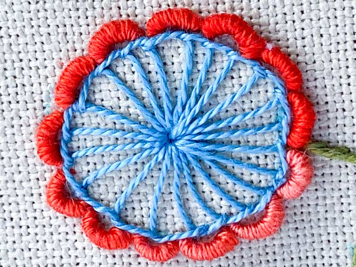 A close-up photo of a flower stitched with blue DMC perle cotton in the center and bullion knots stitched with a salmon-colored DMC floss around the edge to make the flower petals. A small part of the stem is shown on the right side of the photo and the beige Charles Craft Monaco Needlework Fabric is in the background.