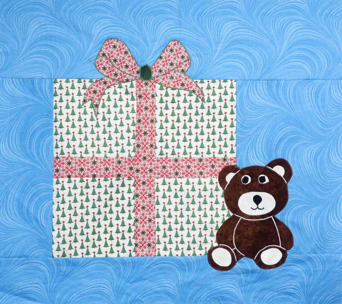 The fabric teddy bear is secured to the Christmas-themed fabric wall hanging using a satin stitch; Odif 808 Spray and Fix Temporary Adhesive for Paper Patterns, Odif 606 Spray and Fix No-sew Fusible Adhesive Web, Odif 404 Spray and Fix Permanent Repositionable Adhesive for Craft Material, Odif 505 Temporary Quilt Basting Adhesive Fabric Spray