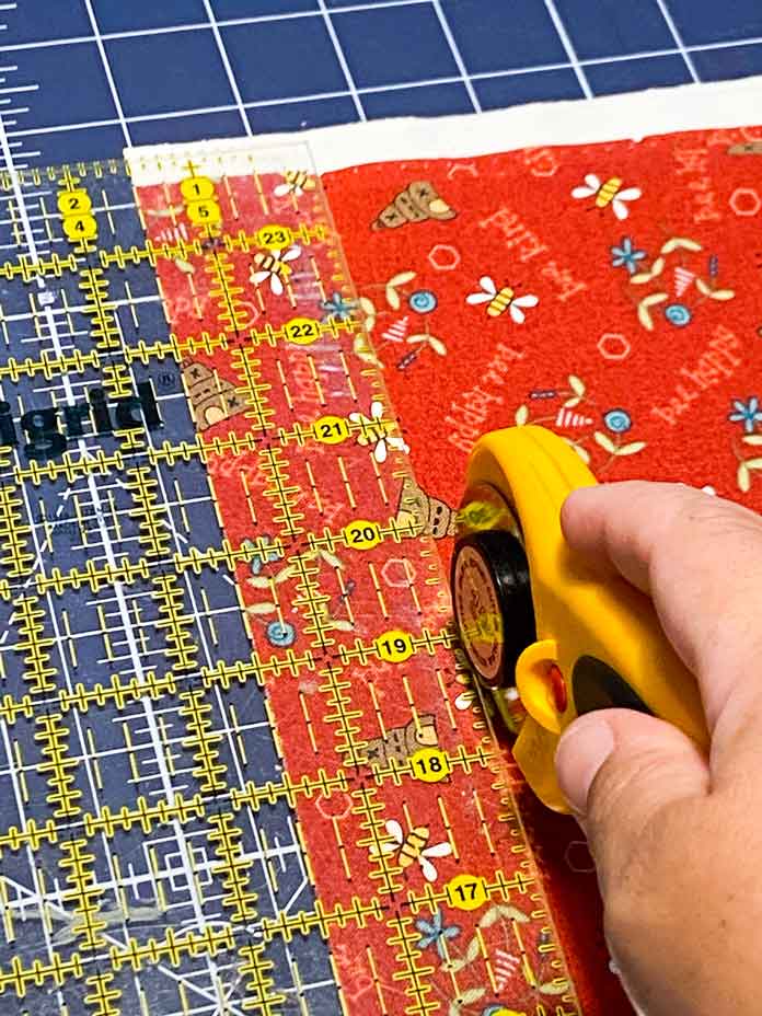 A hand is shown holding a yellow rotary cutter to cut a strip of red fabric using a yellow gridded ruler. A blue rotary cutting mat is shown in the background; Omnigrid ruler - 6" x 24", OLFA Rotary Cutter, OLFA Double Sided Cutting Mat (navy blue)