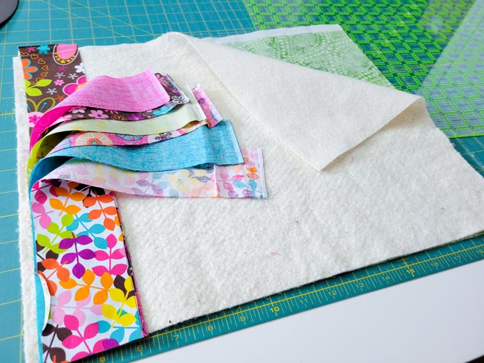 Pink, turquoise, and brown floral fabric strips and a piece of batting laid out on a green cutting mat with a square quilting ruler on the top right corner